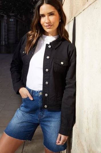 A woman wears a pair of blue Bermuda denim shorts, a white t-shirt and a black cardigan with silver buttons.