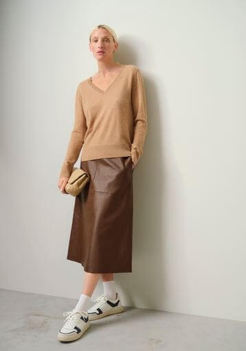 A model wears a pair of white and black trainers with a brown leather midi skirt and a brown jumper.