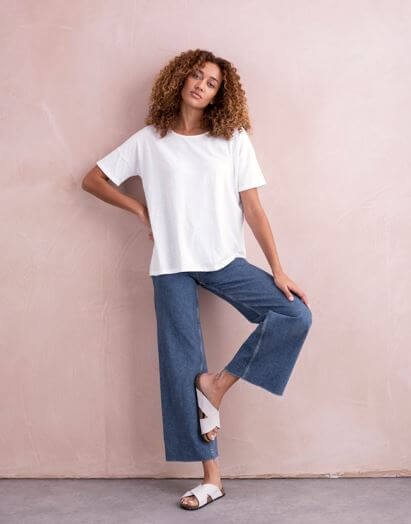A woman with brown curly hair wears a white t-shirt and cropped blue jeans and sandals.