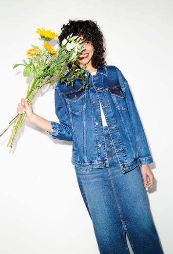 A woman with black curly hair wears a denim jacket and denim skirt.
