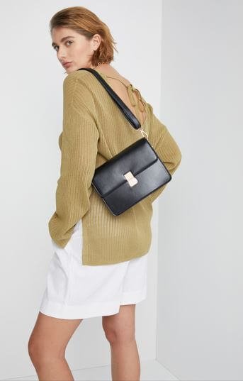 A model wears a black leather cross body bag with white shorts and an olive coloured jumper.