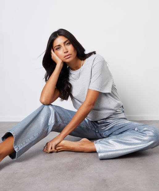 A woman wears a grey t-shirt and a pair of silver metallic denim trousers.