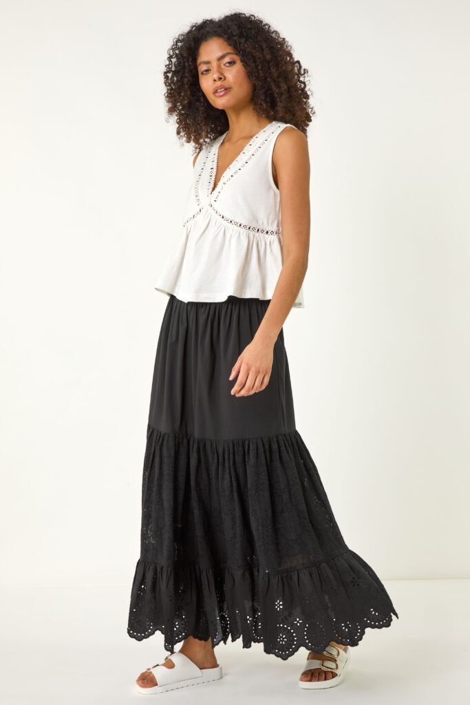 A woman wears a black tired maxi skirt, a white flowy vest top and white flat sandals.
