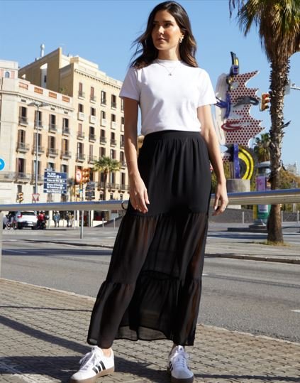 A woman wears a long black maxi skirt, white t-shirt and white Adidas trainers
