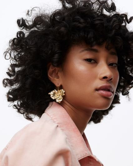 A model with dark curly hair wears statement gold earrings in the shape of a flower.