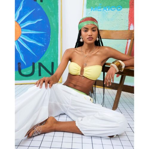 A model wears a boho beach outfit with white cargo pants, yellow bikini top, and bandana.
