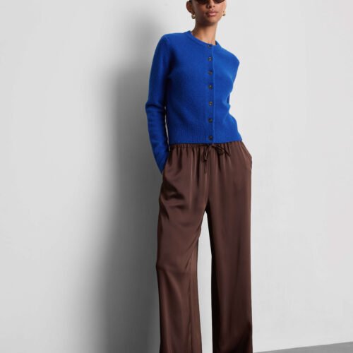 A model wears a bright blue crew neck cardigan, brown wide-leg trousers, brown loafers, brown sunglasses gold hoop earrings.
