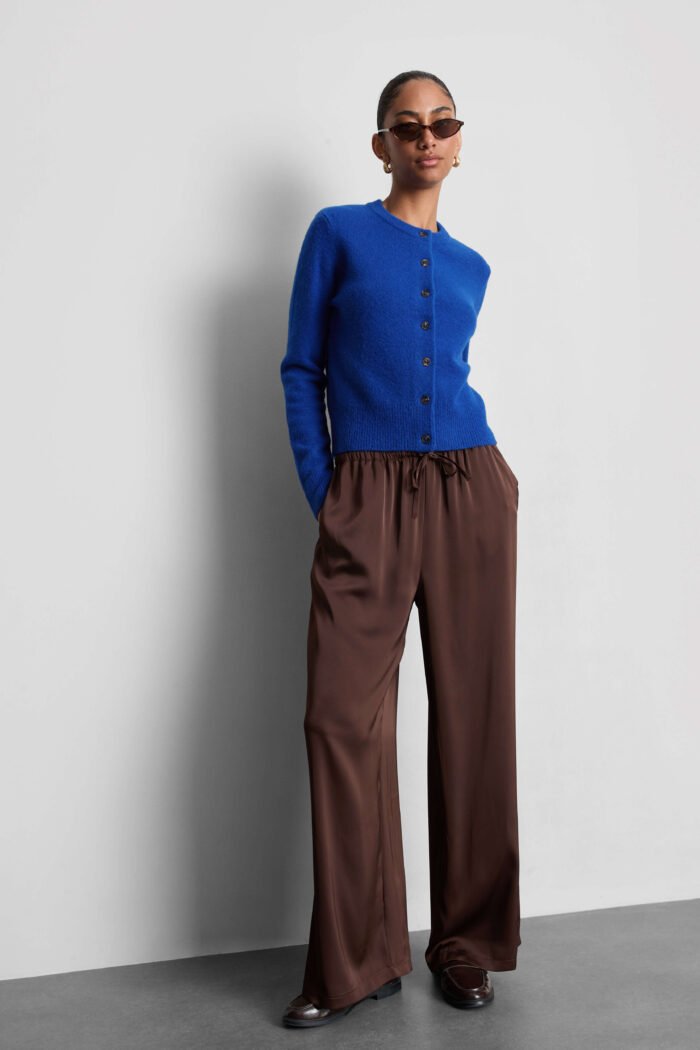 A model wears a bright blue crew neck cardigan, brown wide-leg trousers, brown loafers, brown sunglasses gold hoop earrings.