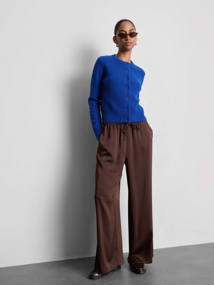 A model wears a bright blue crew neck cardigan, brown wide-leg trousers, brown loafers, brown sunglasses gold hoop earrings.