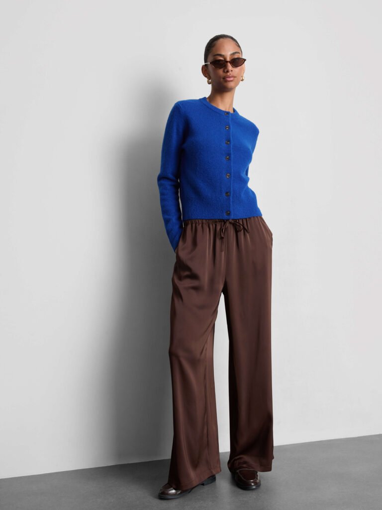 A model wears a bright blue crew neck cardigan, brown wide-leg trousers, brown loafers, brown sunglasses gold hoop earrings.